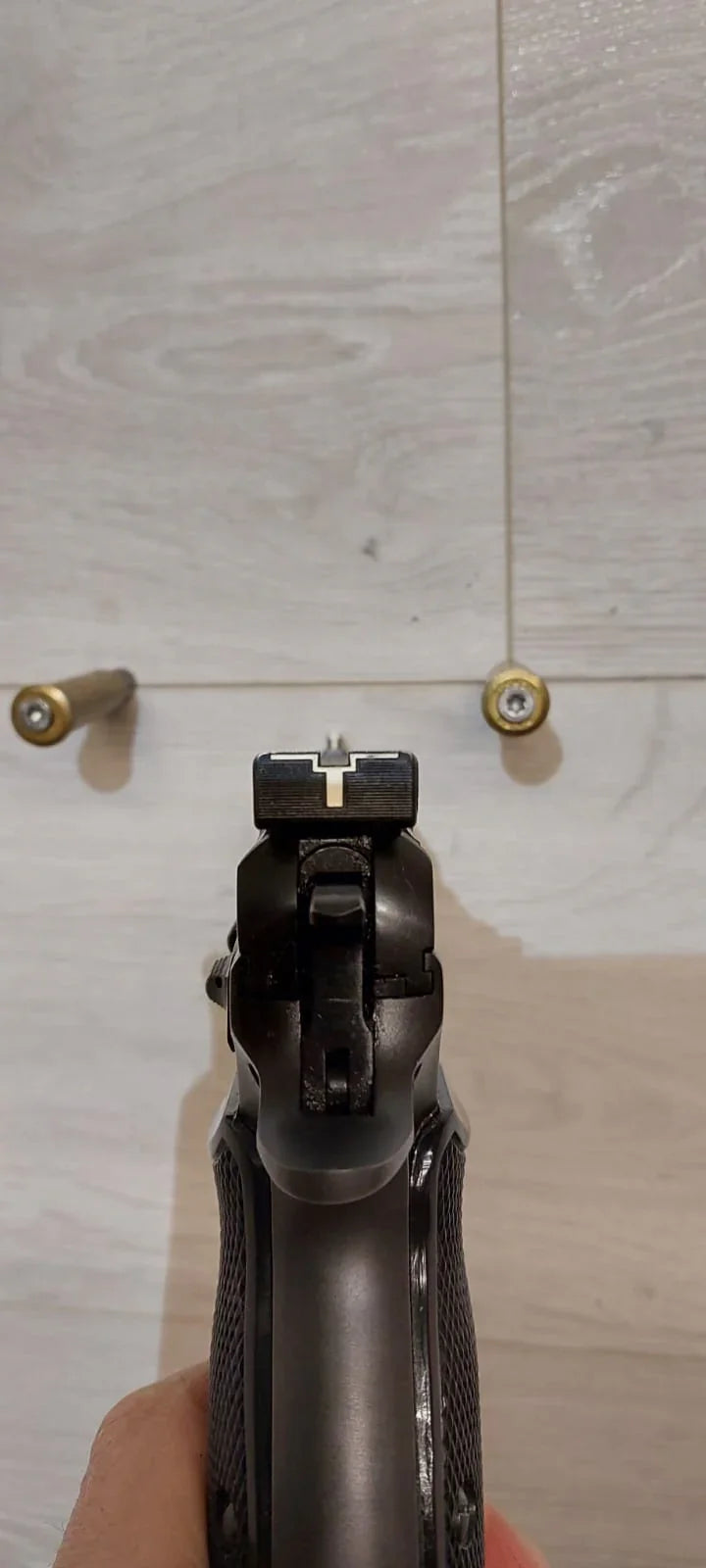Black handgun with textured grip and rear sight, held against light wood background with two brass cartridges.