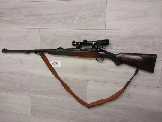 Bolt-action rifle with scope and leather sling on wood floor, ideal for hunting or sport shooting.