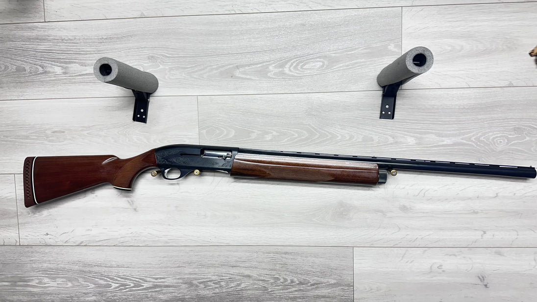 Elegant wooden stock shotgun with engraved receiver, displayed on light wood floor with foam gun rests.