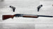 Elegant wooden stock shotgun with engraved receiver, displayed on light wood floor with foam gun rests.