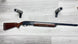 Elegant wooden stock shotgun with engraved receiver, displayed on light wood floor with foam gun rests.