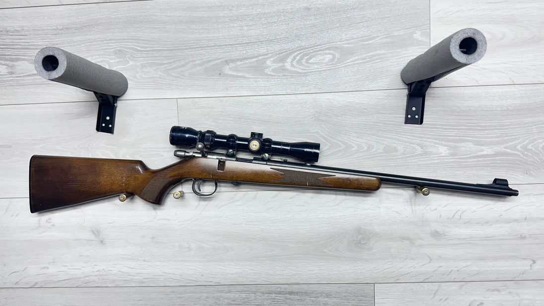 Wooden stock rifle with scope and ammo, displayed on light wood floor with foam gun rests