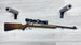 Wooden stock rifle with scope and ammo, displayed on light wood floor with foam gun rests
