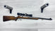 Wooden stock rifle with scope and ammo, displayed on light wood floor with foam gun rests