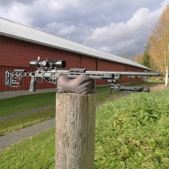 Precision rifle with scope on wooden post, ideal for competition shooting and hunting, set outdoors near red barn.