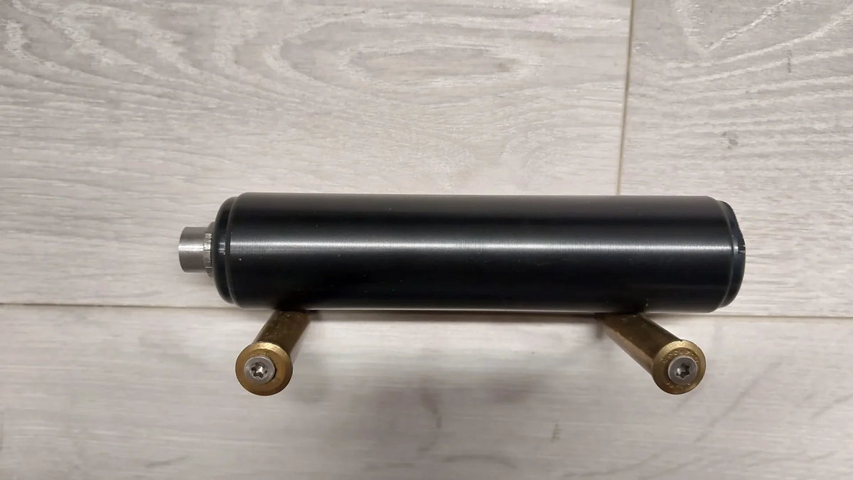 Black cylindrical firearm component with brass cartridge mounts on light wood surface