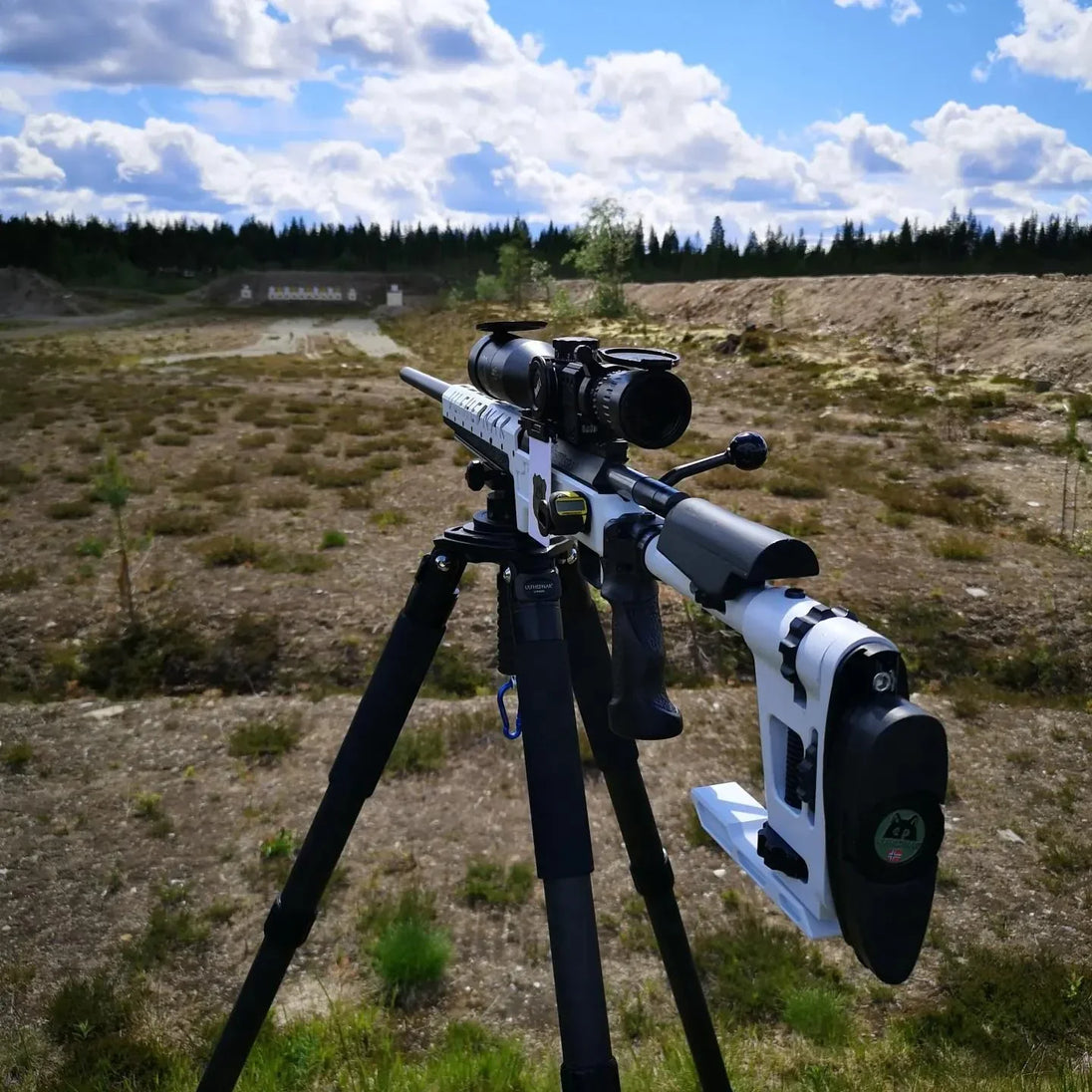 White precision rifle with scope on tripod at outdoor shooting range under cloudy sky