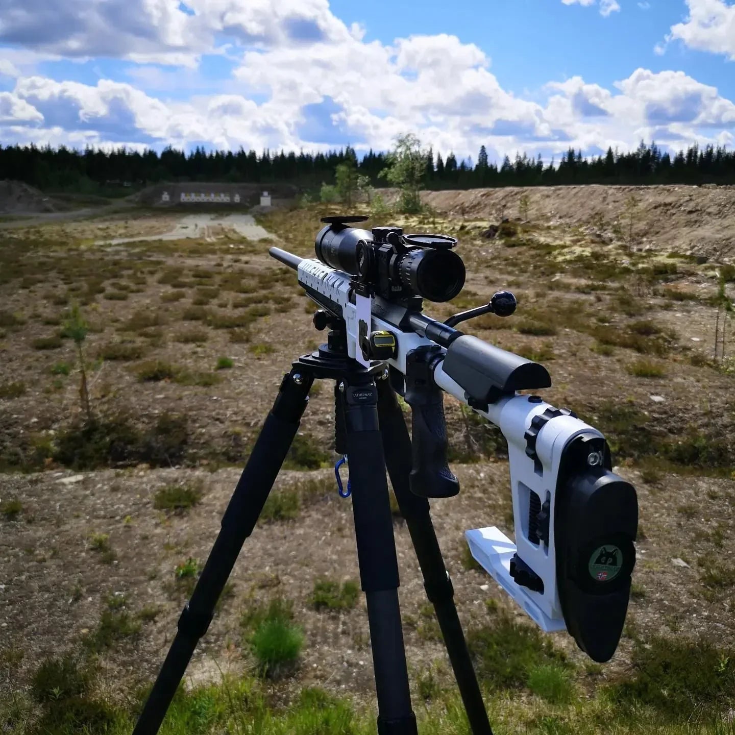 White precision rifle with scope on tripod at outdoor shooting range under cloudy sky