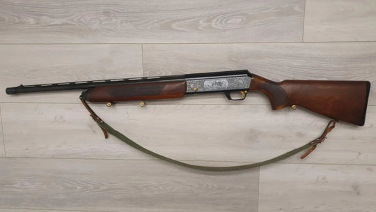 Vintage hunting shotgun with engraved receiver, wooden stock, and green sling on wood floor
